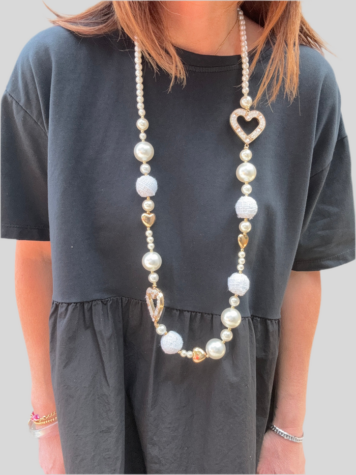 Collier long fantaisie composé de perles nacrées, cœurs dorés et boules tissées blanches, avec pendentif cœur strassé. Look romantique et lumineux porté sur une robe noire.