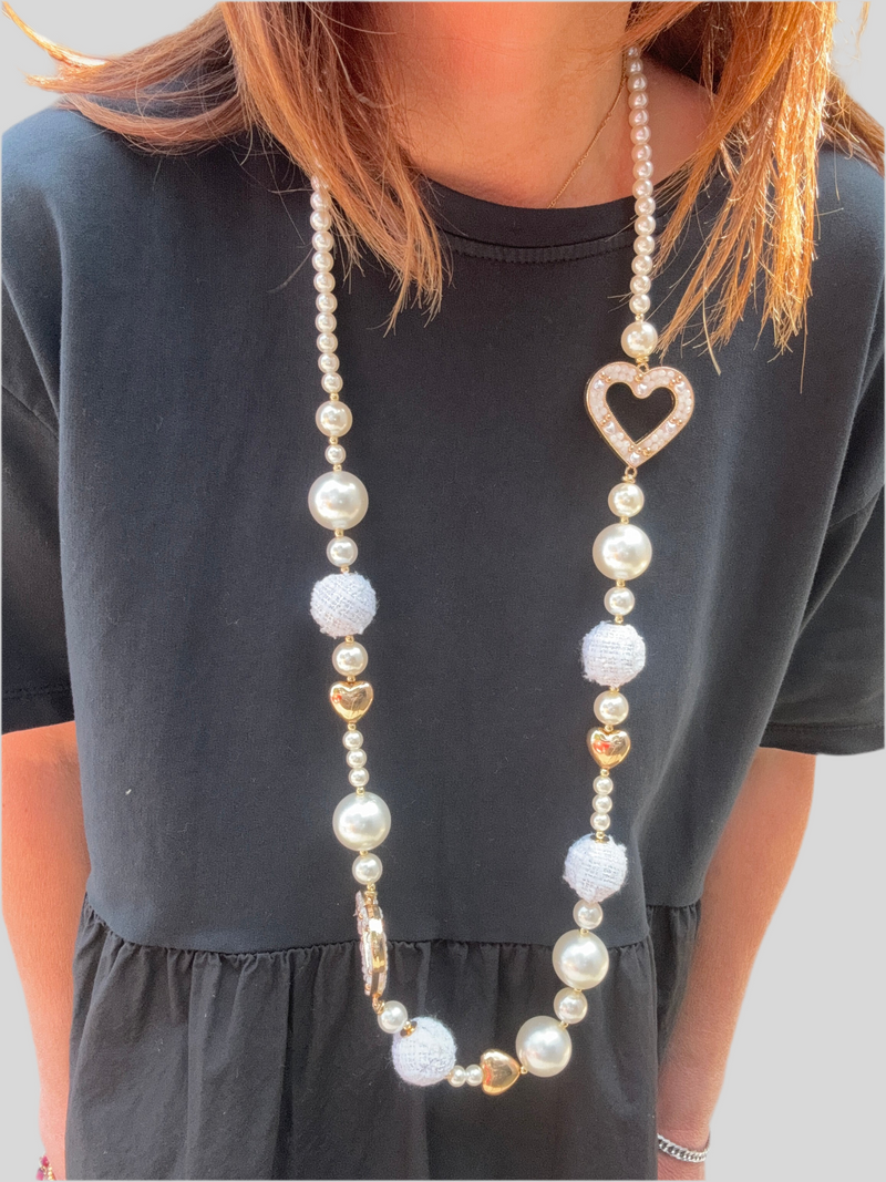 Collier long fantaisie composé de perles nacrées, cœurs dorés et boules tissées blanches, avec pendentif cœur strassé. Look romantique et lumineux porté sur une robe noire.