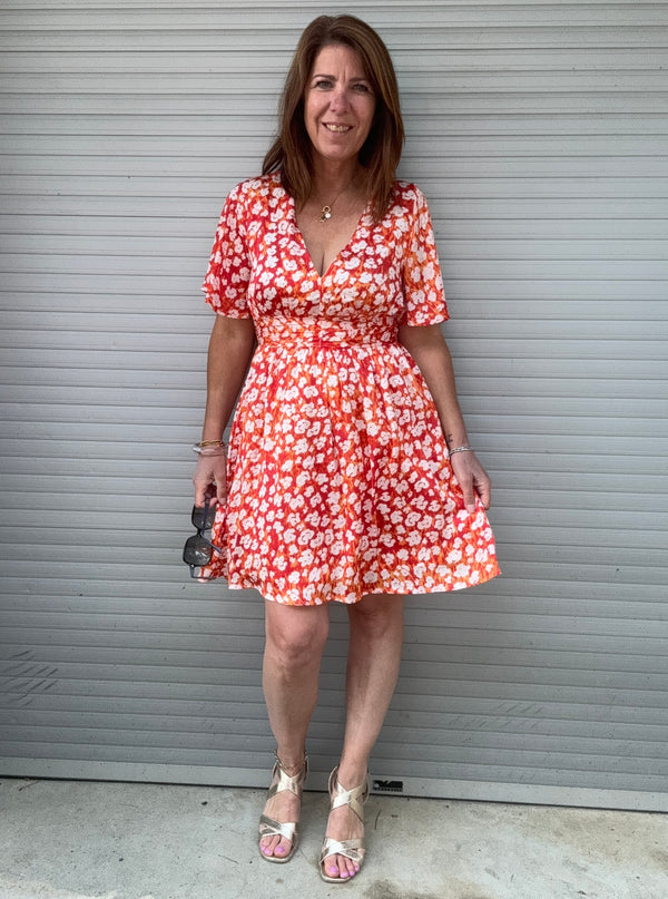 Robe courte à fleurs rouges, orange et blanches portée par une femme, coupe patineuse, manches courtes, décolleté cache-cœur. Taille marquée et doublure intégrale. Look estival féminin.charly 69390